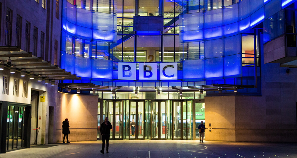 View of the BBC corporate building at night with blue lights and people walking thru the entrance.